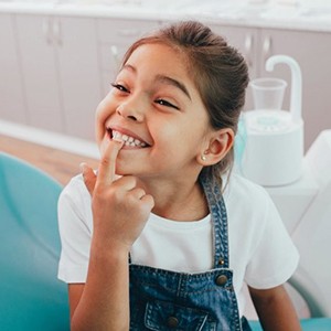 little girl sitting in dental chair and pointing to her teeth 